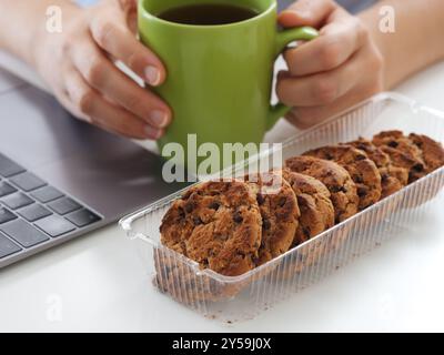 Businesswoman sitting behind a laptop, holding a coffee cup and getting ready to eat chocolate chip cookies during a break at work. Close up. Stock Photo