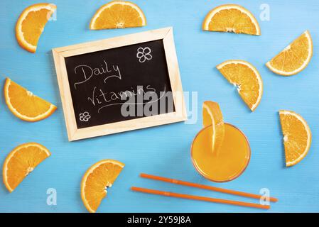 High angle image of a blackboard with the message daily vitamins, surrounded by slices of orange, a glass of juice and straws, on a blue background Stock Photo