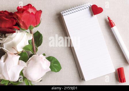 Open spiral notebook with stripes and a wooden clip with red heart pinned on it and a bouquet of white and red roses, on a vintage fabric background Stock Photo