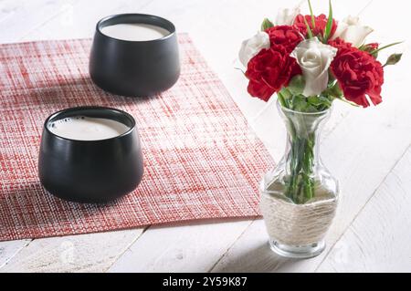 Breakfast table with two black mugs full of fresh milk and a bouquet of white roses and red carnations, on a white table, on a sunny morning Stock Photo