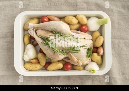 Roast chicken prepared for roasting with young garlic, shallots, parsley, cherry tomatoes, potatoes and olive oil in a casserole dish Stock Photo