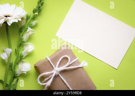 Greeting card idea with a paper note surrounded by white flowers and a gift box wrapped in brown paper and tied with flax string and bow, on a green b Stock Photo