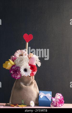 Bouquet of roses, gerbera, tulips and a red heart, in a jute bag, surrounded flowers and a cute blue gift box, with a black vintage background Stock Photo