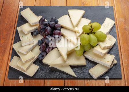 Slices of brie and toma cheese with clusters of red and white grapes on black slate plate isolated on on wooden table, clipping path included Stock Photo