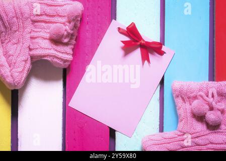 Unwritten pink paper note with red bow surrounded by handmade knitted pink bootees, on a multicolored wooden background Stock Photo