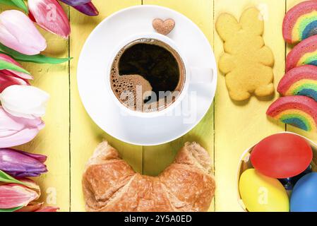 Easter morning with a cup of coffee, rainbow cookies, painted eggs, bunny shaped cookie, croissant and multicolored tulips, on a yellow wooden table Stock Photo