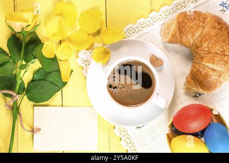 Breakfast table with a cup of coffee, freshly baked croissant, painted Easter eggs and a yellow single rose with fallen petals and a message card Stock Photo