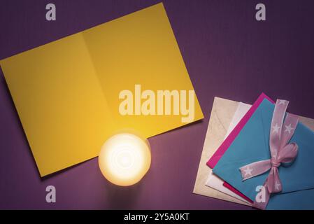 Empty paper message card, a lit candle on it and a pile of multicolored envelopes tied with ribbon and bow, on a purple wooden background Stock Photo