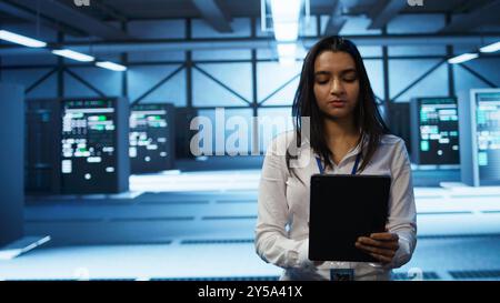 Software developer walking in data center, using tablet to design, implement and manage gear, ensuring optimal performance and reliability. IT worker in server farm coding on device, camera B Stock Photo