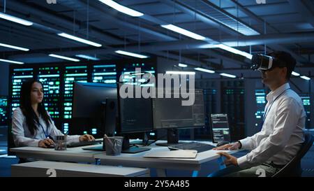 Admin in data center using virtual reality tech to ensure system can handle networking tasks. Server room worker wearing VR headset to track network traffic with PC, making improvements, camera B Stock Photo