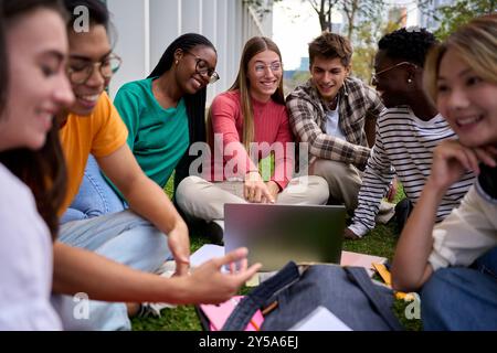 Group diverse Gen z university joyful students going through syllabus sitting on lawn in campus park Stock Photo