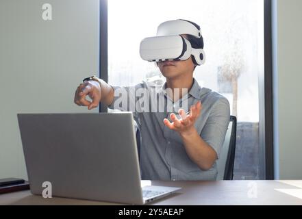 At office, Using VR headset, asian man interacting with virtual environment while working on laptop Stock Photo