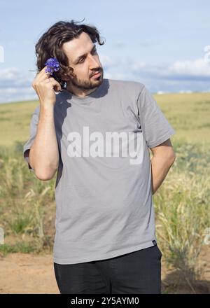 Curly-haired guy in gray shirt with cornflowers on background of wheat field in summer. Sunny day. Mock up for design.  Stock Photo