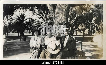 A relaxed trio sits on a park bench beneath the shade of a large tree, enjoying a sunny day surrounded by palm trees. The two women and a man, all dressed in classic early 20th-century attire, pose for the camera in this serene, outdoor setting Stock Photo