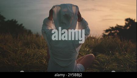 Woman in a white jacket, sitting in a grassy field at sunset, putting on its hood, surrounded by nature's serene atmosphere, perfect for meditation or yoga Stock Photo