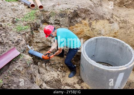 Installing plastic drain underground pipe, gravity drainage, plumber installs socket bend in front of septic tank made of concrete rings. Stock Photo