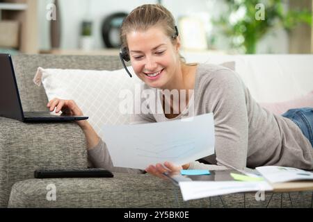 woman lays on sofa wearing headset and using laptop Stock Photo