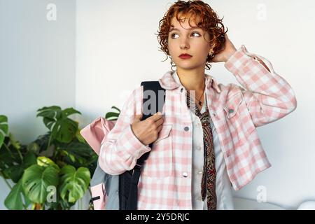 attractive extravagantly dressed young woman, student, schoolgirl with red curly hair with backpack in home interior Stock Photo