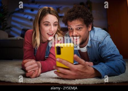 Young Caucasian couple looking with surprised face at phone lying on carpet in living room at night. Stock Photo