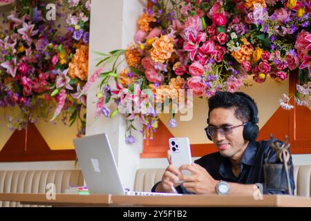 handsome asian male freelancer wearing glasses wearing headphones smiling operating mobile phone in front of laptop computer on table, sitting in indo Stock Photo