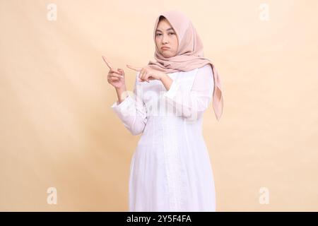 young asian muslim woman standing to camera pouting pointing to top right. Fashion, pointer, business and promotion concept Stock Photo