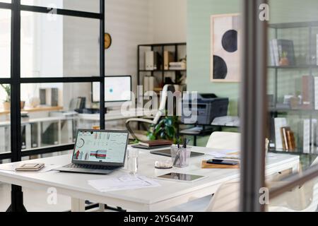 Modern office interior featuring an open workspace with glass partitions, workstations, and greenery enhancing a productive environment. Equipment includes laptops, shelves, and printers Stock Photo