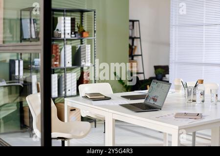 Modern office with white desk featuring open laptop, notebooks, and potted plants on shelves. Sunlight filtering through the blinds illuminating workspace Stock Photo