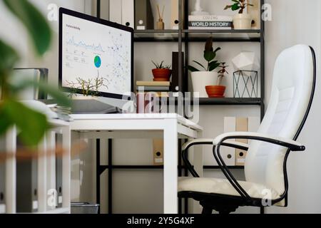 Modern white office space featuring computer monitor, shelves filled with books and plants, and comfortable chair inviting organized work atmosphere Stock Photo