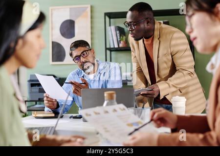 Group of professionals collaborating on business strategy in modern office environment with documents, devices, and focused expressions on their faces creating productive atmosphere Stock Photo