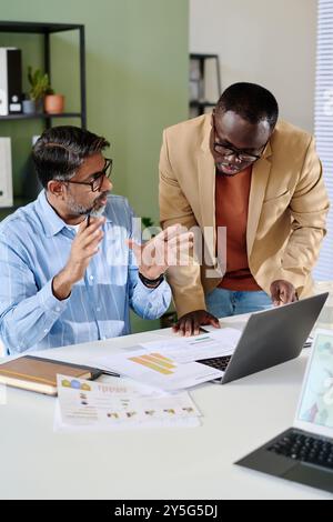 Two professionals discussing project while reviewing documents and using laptop in office environment displaying teamwork and collaboration Stock Photo