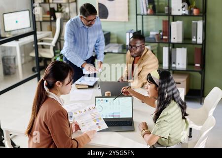 Group of co-workers interacting in modern office with open laptops on table, discussing project details. Diverse team utilizing documents and charts during work meeting Stock Photo
