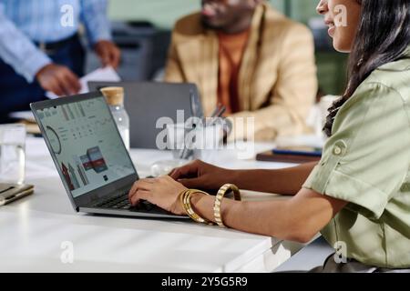 Man and woman collaborating in office analyzing business data on laptop screen with charts and graphs surrounding them. Visible documents and coffee cup on table enhancing professional atmosphere Stock Photo