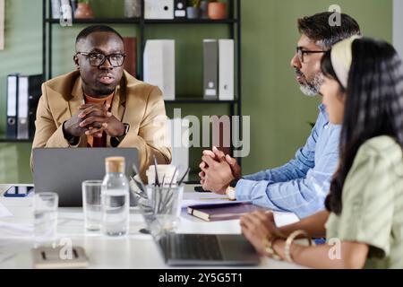 Three professionals discussing business strategies around an office table with laptops and documents. Man in brown blazer leading brainstorming session, engaging team members Stock Photo