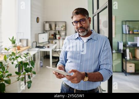 Portrait of bearded man in blue shirt holding tablet while standing in modern office setting with shelves and plants, creating an inviting and professional atmosphere Stock Photo