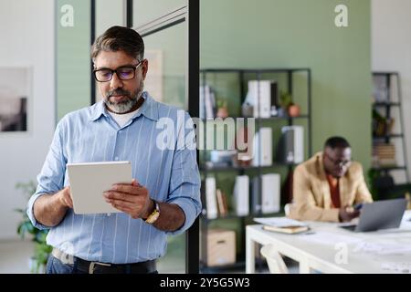 Hispanic senior man using tablet while leaning against glass wall in modern office environment while another man sits at desk in background working on laptop Stock Photo