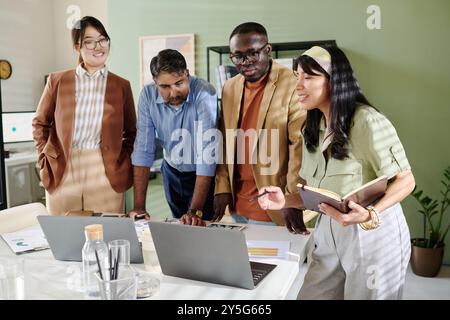 Group of diverse professionals collaborating on project while standing around table with laptops and documents looking engaged in discussion in modern office environment Stock Photo