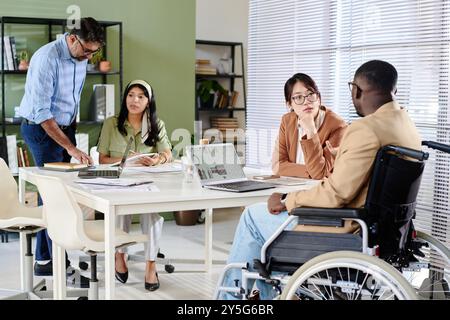Group of colleagues working together in office, including person in wheelchair, engaging in discussion around laptops on table during meeting Stock Photo