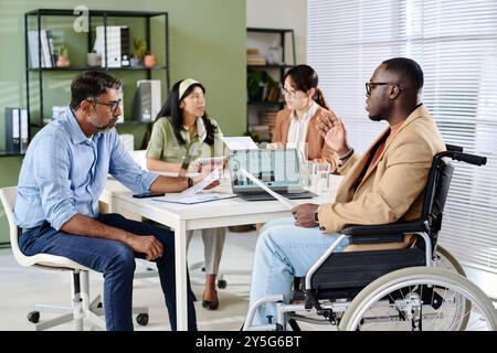 Group sitting around table in office discussing strategies while one man in wheelchair presenting data using laptop engaging team members in conversation Stock Photo
