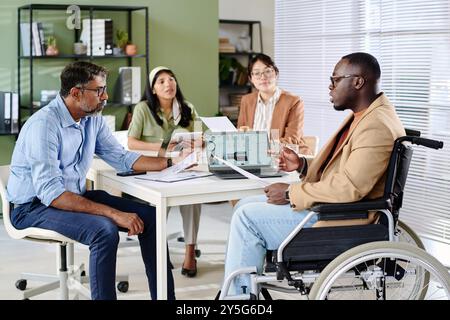 Group of business professionals sitting around table, engaging in important meeting. People reviewing documents, one person in wheelchair, modern office environment Stock Photo