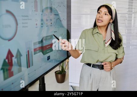 Woman wearing headscarf presenting data on large screen in office setting Holding pointer, she is discussing financial and analytical growth charts indoors, possibly during business meeting Stock Photo