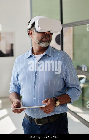 Man engaged in virtual reality experience while holding tablet in modern office environment utilizing VR headset wearing casual blue shirt and standing near a window Stock Photo