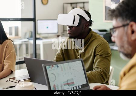 Man immersed in virtual reality using VR headset while sitting in contemporary office space alongside two colleagues collaborating on digital devices Stock Photo