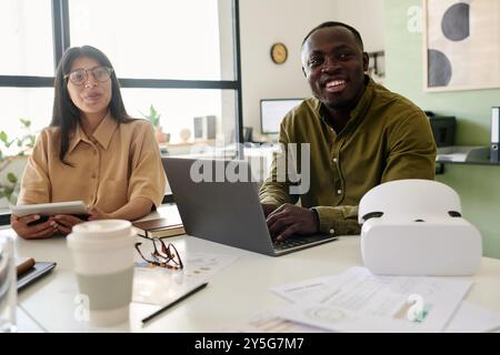 Two colleagues, one Black person and one Caucasian, smiling and collaborating on project with laptop and documents on table, enjoying bright and open workspace Stock Photo