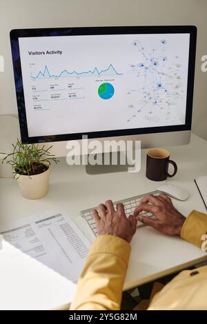 Image shows person analyzing visitors activity data on large computer screen with charts and graphs, coffee mug, and plant on desk Stock Photo