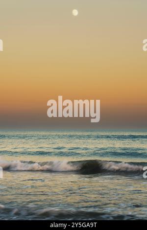 Calm waves roll toward the shore beneath a pale moon and pastel sky. Stock Photo