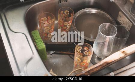 Untidy metal sink full of dirty dishes and tableware. Pile of dirty washing up utensil in the kitchen. Stock Photo