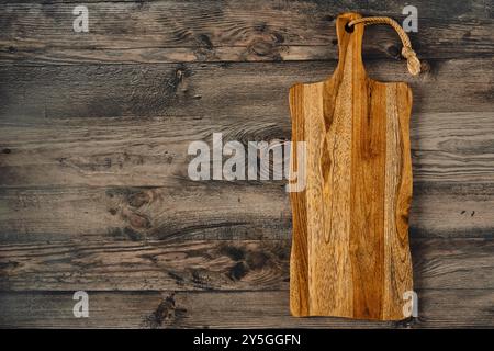 A handcrafted wooden cutting board rests on a weathered wooden table, displaying rich grains and unique patterns. The board features a rope handle, em Stock Photo