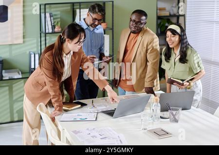 Group of diverse colleagues engaging in collaborative discussion around office table, surrounded by laptops, documents, and stationary, planners examining financial reports together Stock Photo