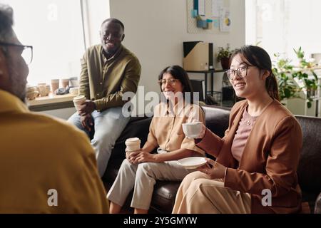 Group of diverse colleagues sitting on couch, smiling and holding coffee cups in bright office setting, creating positive and relaxed atmosphere Stock Photo