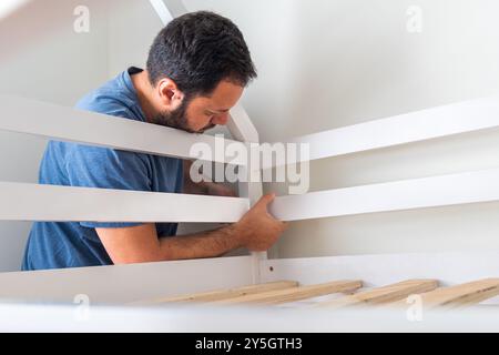 A man is fixing a bed frame. The bed is white and the frame is made of wood Stock Photo